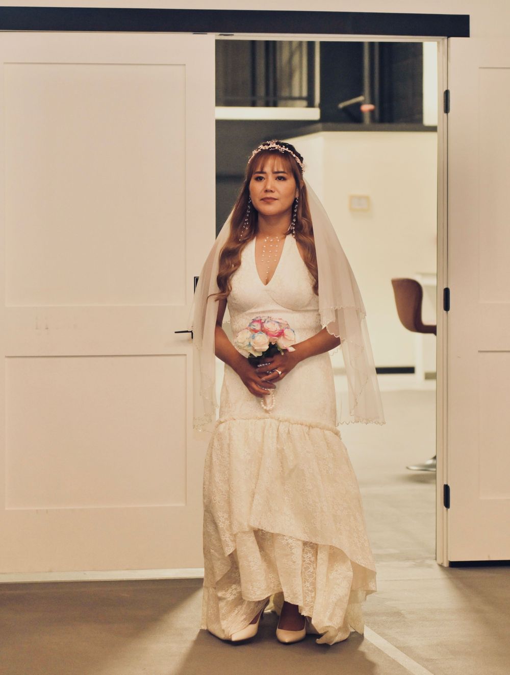 Bride in white lace gown and veil holding pink bouquet standing in doorway.
