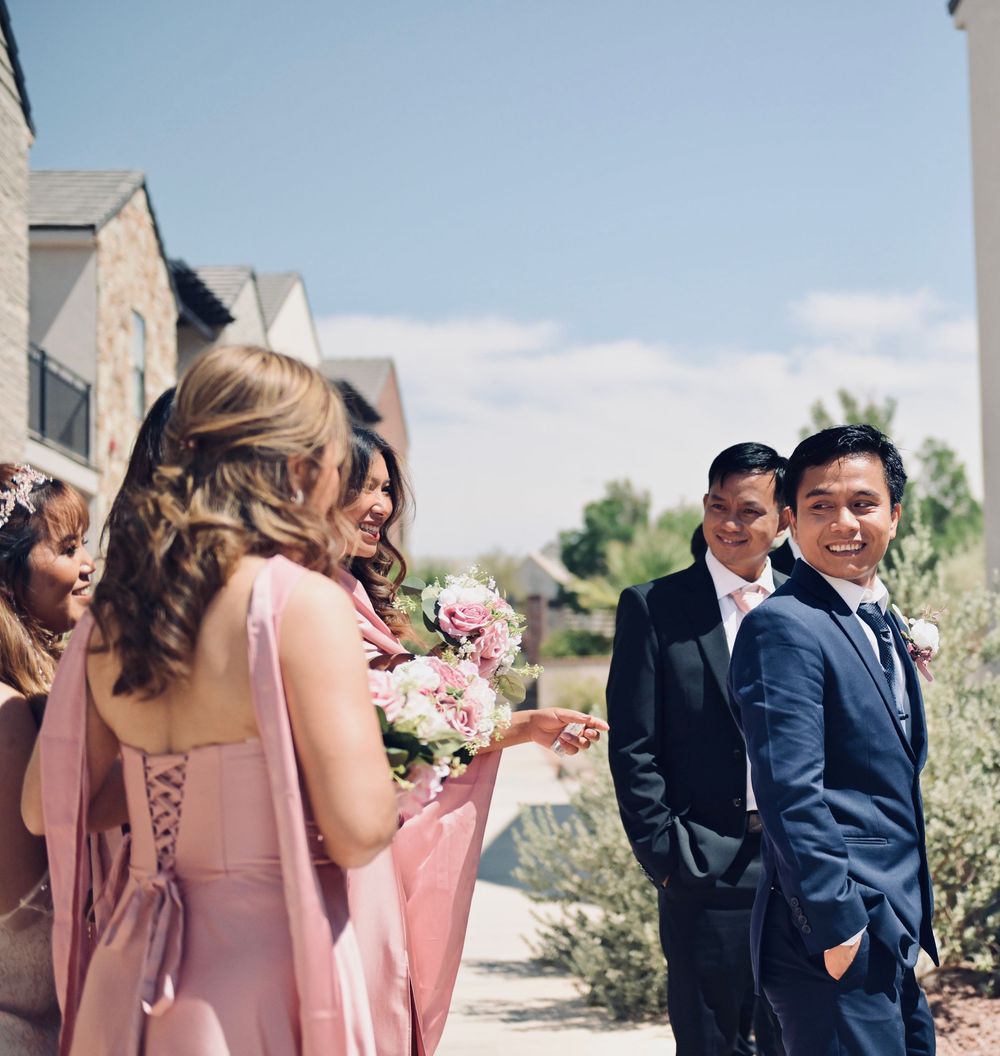Bridesmaids in pink dresses holding bouquets chat outdoors while groomsmen in navy suits stand nearby at a sunny wedding.