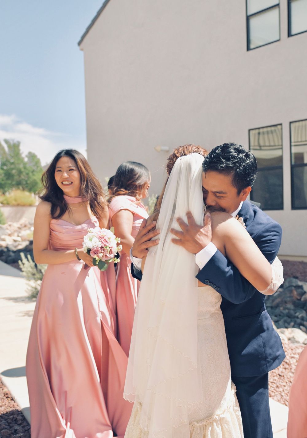 Groom embraces bride in wedding veil outdoors while smiling bridesmaids in pink gowns celebrate nearby.