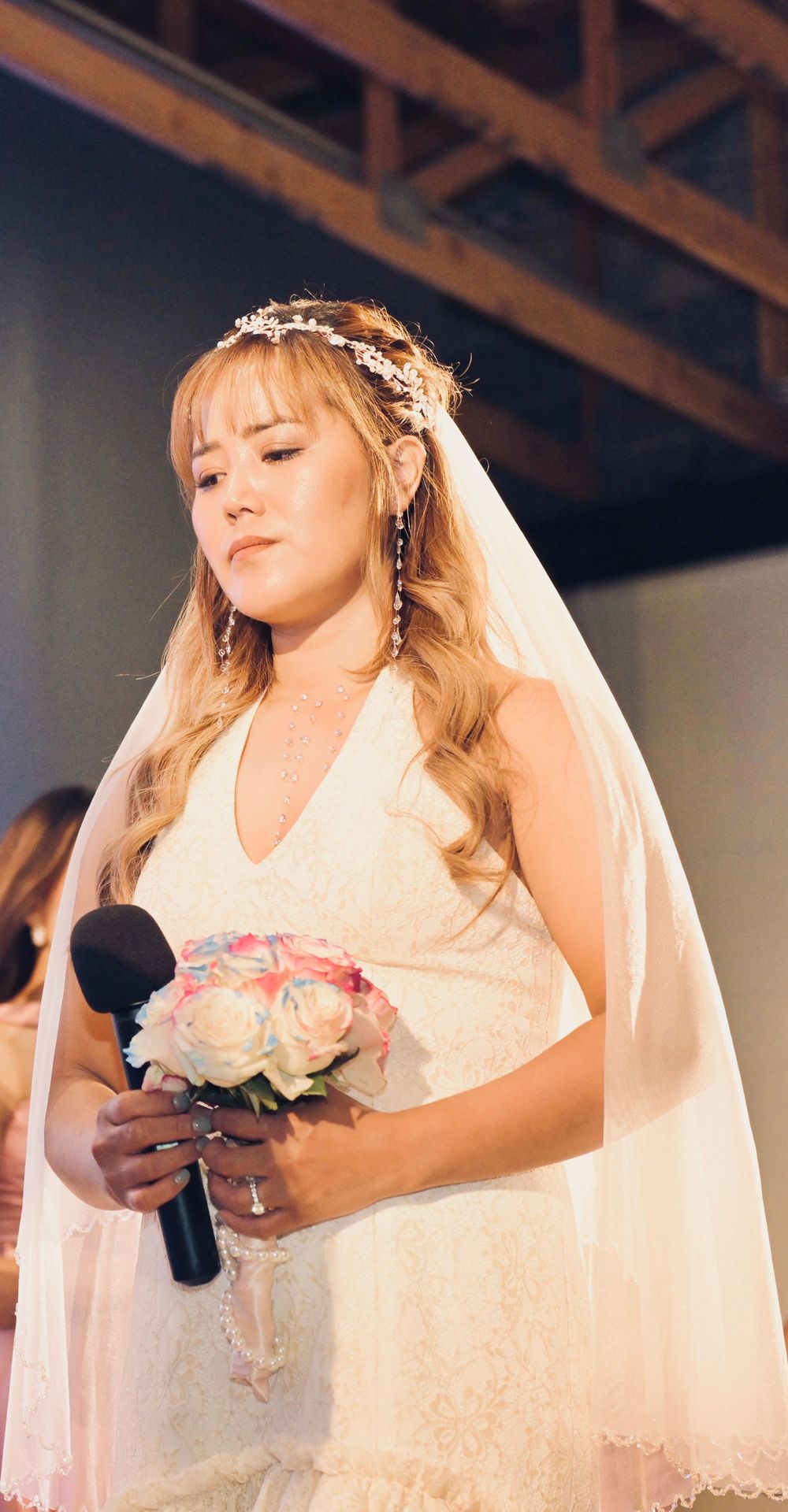 Bride in white lace gown and floral veil holds bouquet and microphone during wedding ceremony speech.