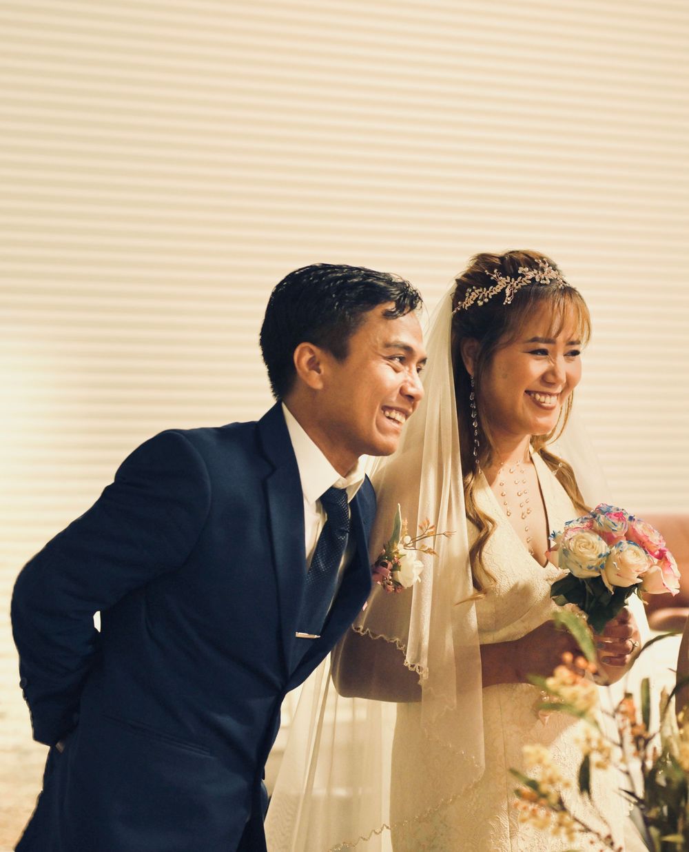 Smiling Asian bride and groom in wedding attire, bride holding bouquet, groom in navy suit.