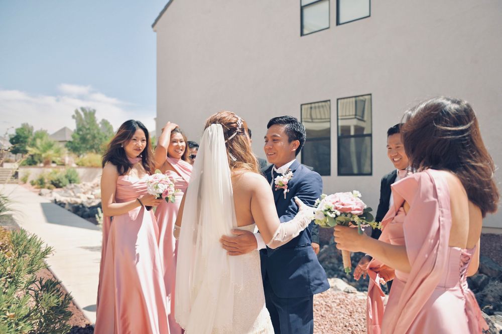 Groom embracing bride in white veil surrounded by bridesmaids in pink dresses at outdoor wedding ceremony.