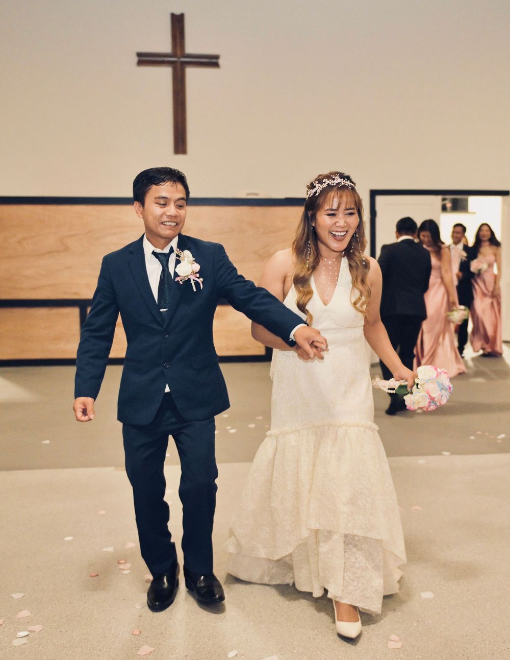 Joyful newlyweds walk hand in hand down church aisle after wedding ceremony beneath wooden cross.