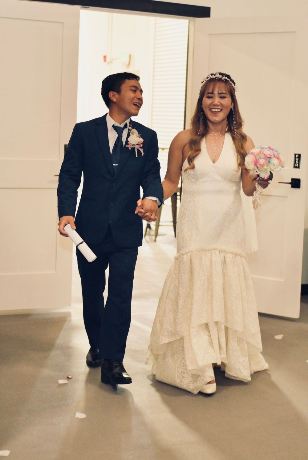 Happy newlyweds walk hand in hand, bride in white gown with bouquet, groom in navy suit after wedding.