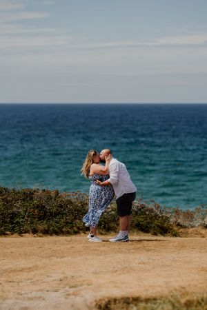 Couple kissing on a coastal clifftop with turquoise ocean in the background on a sunny day.