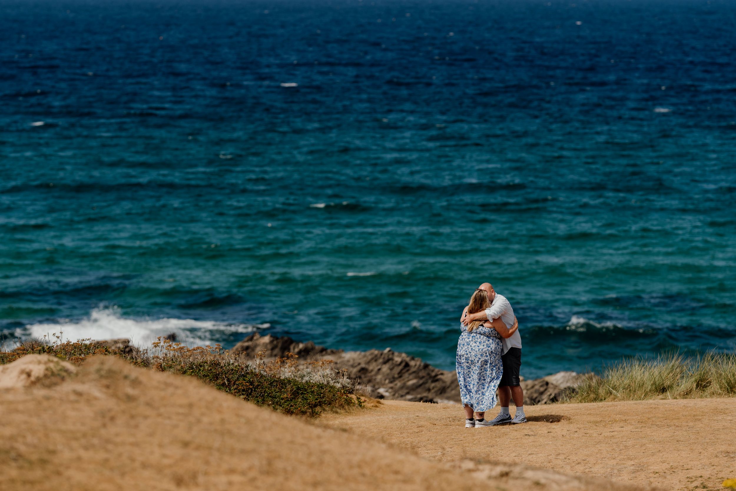 Couple embracing on a coastal cliff overlooking vibrant turquoise ocean waters on a sunny day.