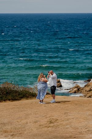 Couple standing on coastal cliff overlooking turquoise ocean waves, man photographing scenic seascape on sunny day.