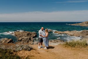 Couple holding hands on rocky coastal path with turquoise ocean and blue sky in background on sunny day.