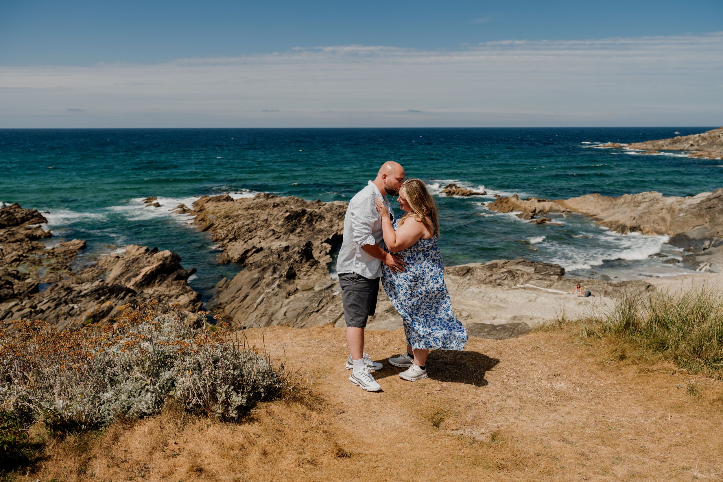 Couple embracing and kissing on rocky coastal path with turquoise ocean waves and blue sky behind them.
