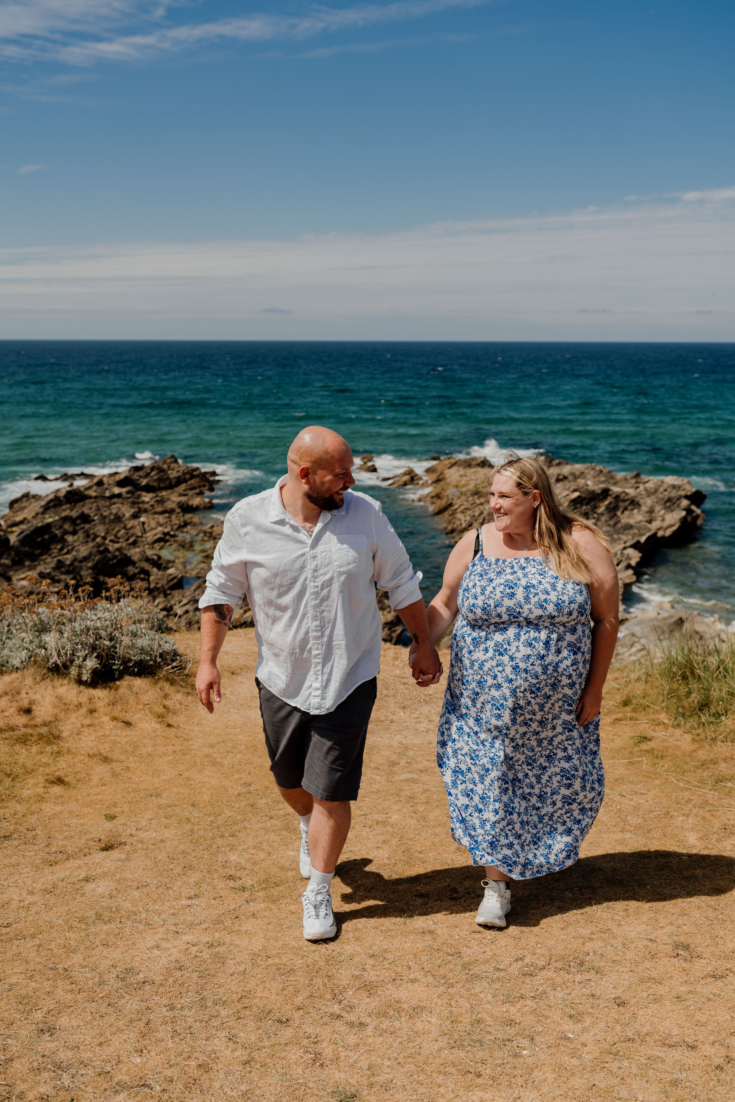 Couple holding hands walking along a coastal cliff path with turquoise ocean views on a sunny day.