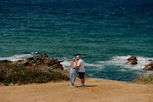 Couple embracing on a rocky coastal cliff overlooking turquoise ocean waves on a sunny day.