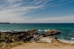 Couple embracing on rocky coastal path with turquoise ocean and blue sky in background.