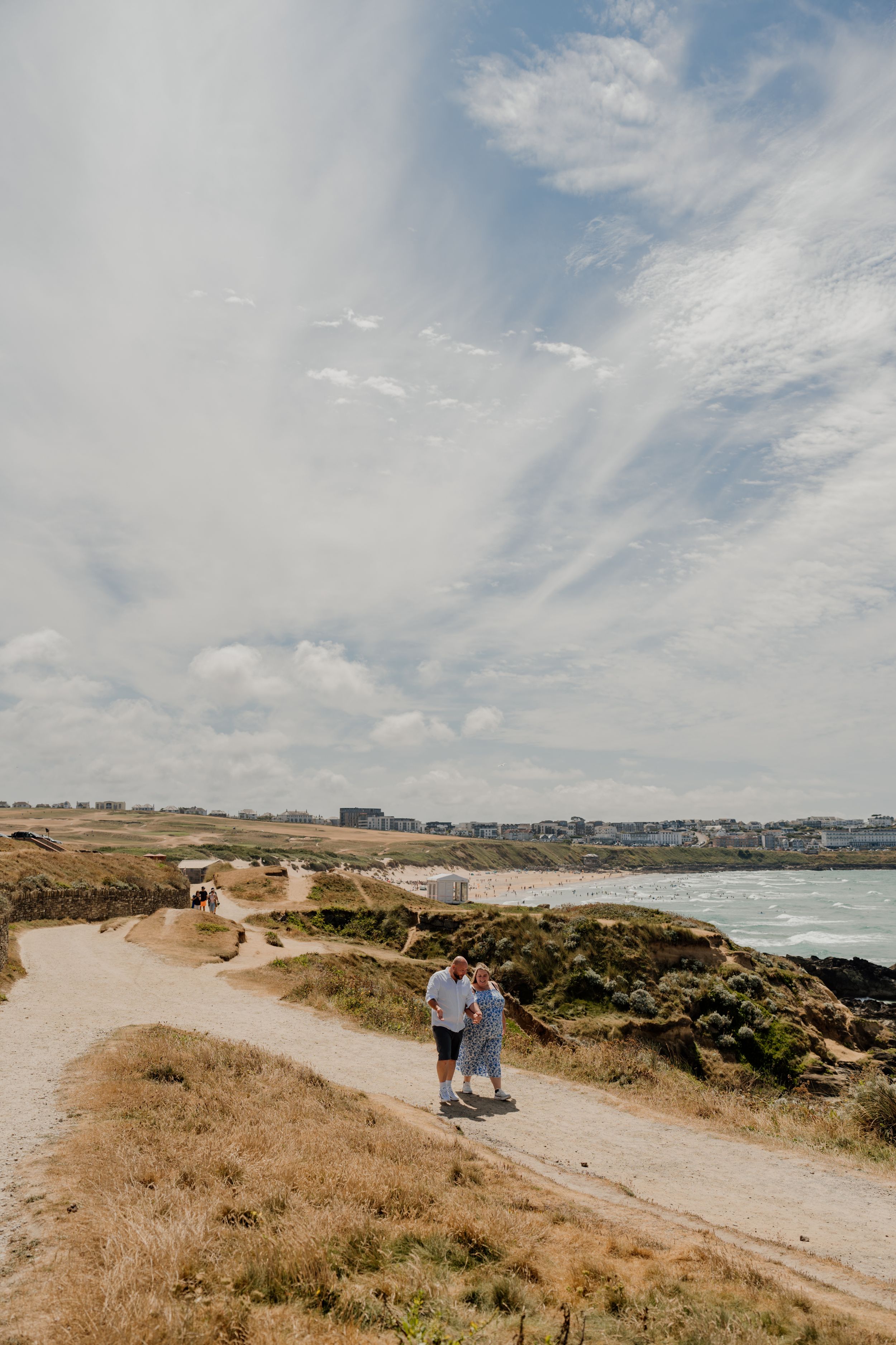 Couple walking on a coastal path with ocean views and cloudy skies in the background.