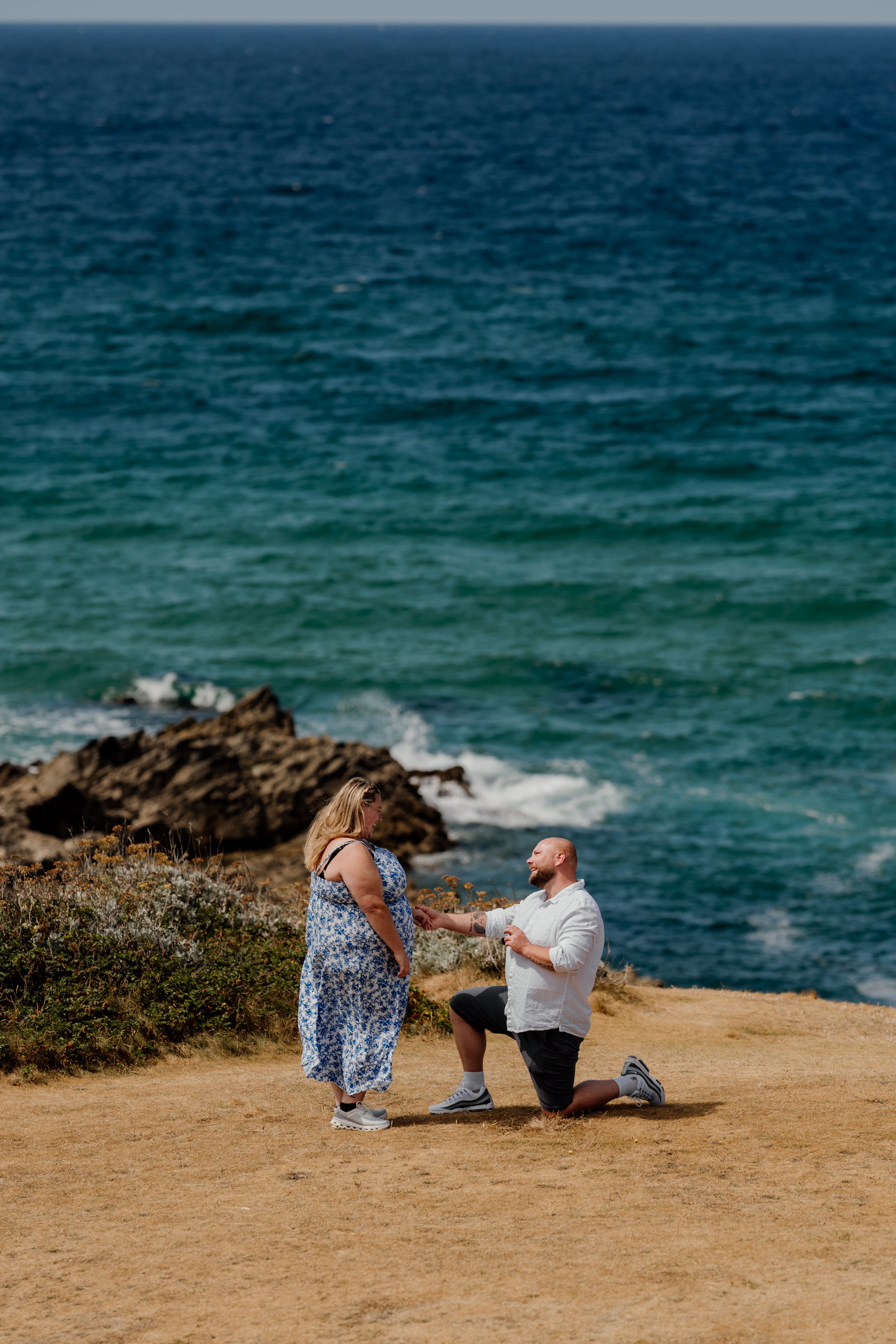 Man proposes on one knee to woman on coastal cliff overlooking turquoise ocean waves and rocky shoreline.