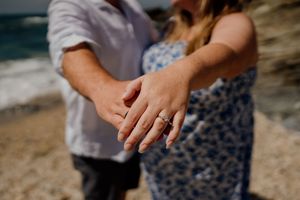 Couple holding hands on a beach showing off an engagement ring, with ocean waves in the background.