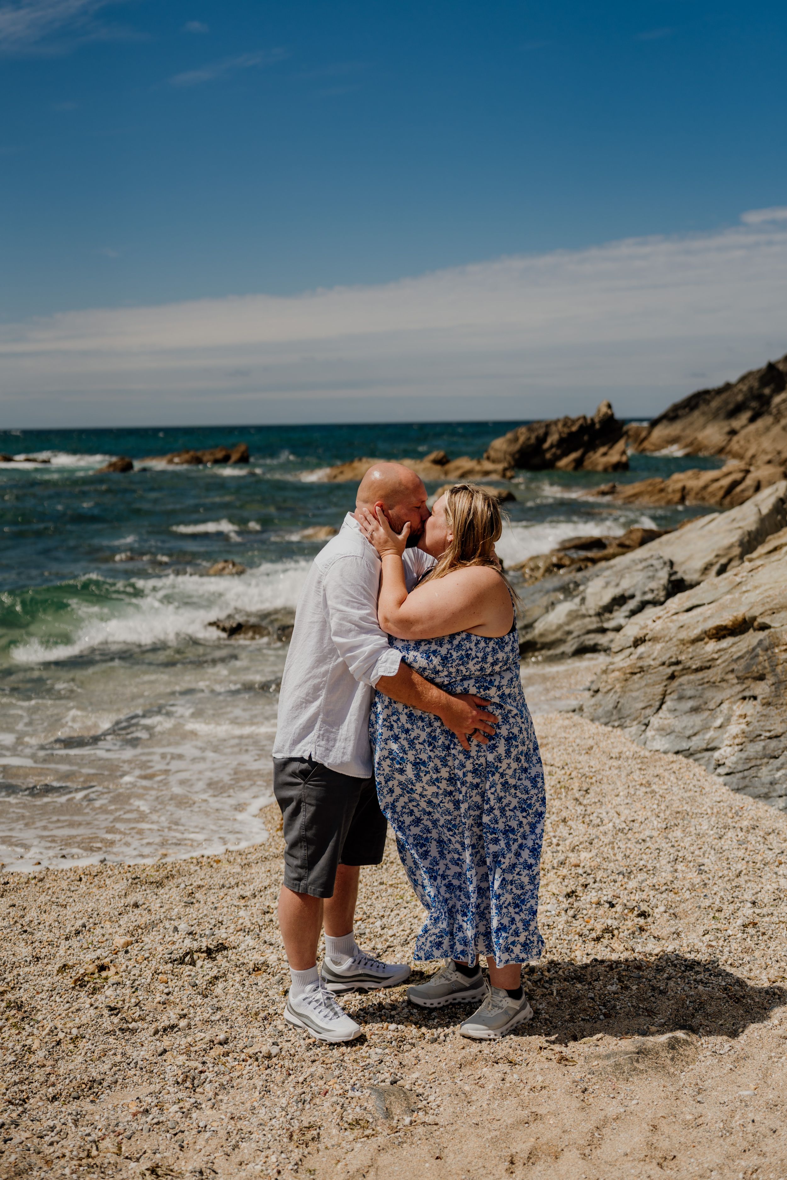 A couple shares a romantic kiss on a rocky beach with ocean waves and blue sky behind them.