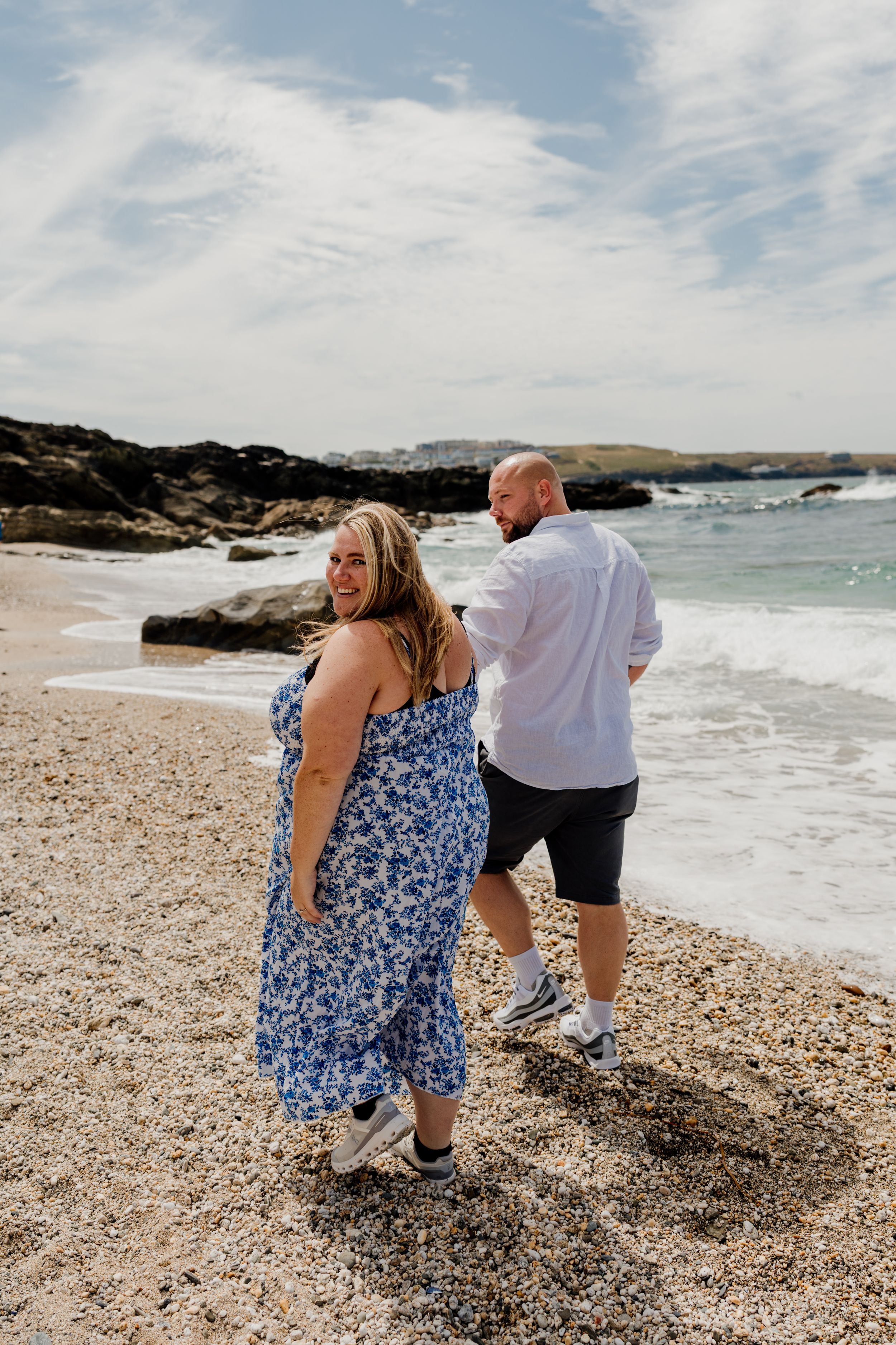 Couple walking on a rocky beach, woman in blue floral dress laughing, man in white shirt and black shorts.