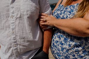 Pregnant woman in blue floral dress holds hands with partner in white linen shirt, showcasing engagement ring.