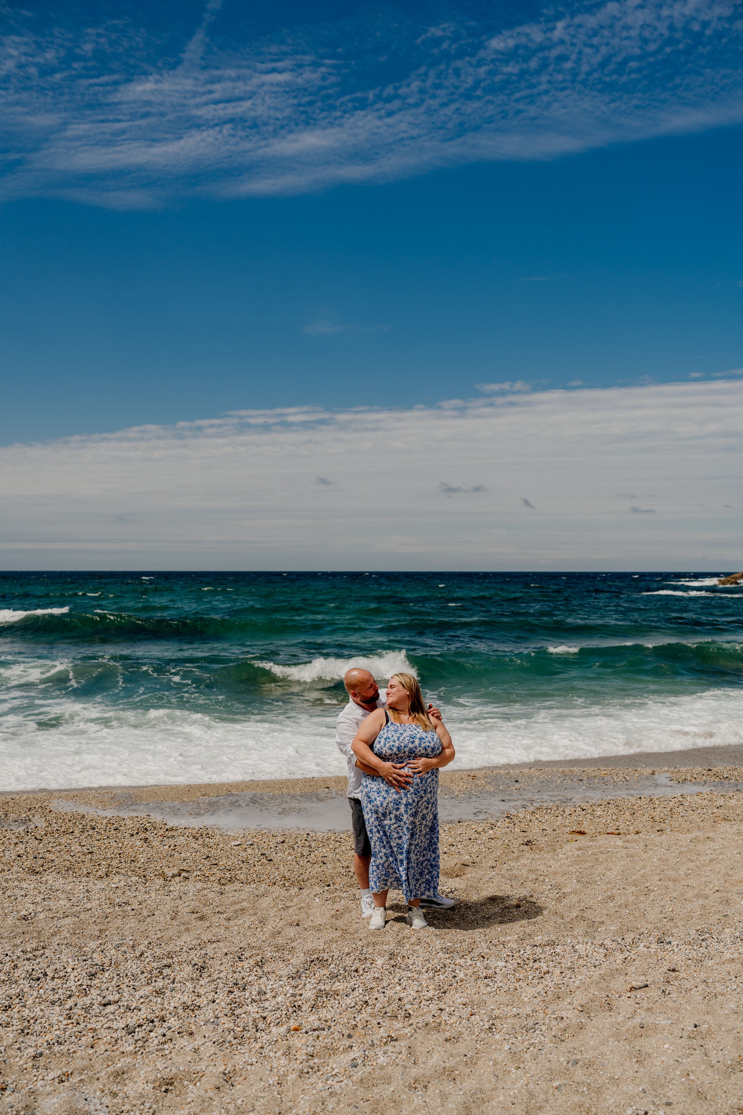 Couple embracing on a sandy beach with turquoise ocean waves and blue sky backdrop.