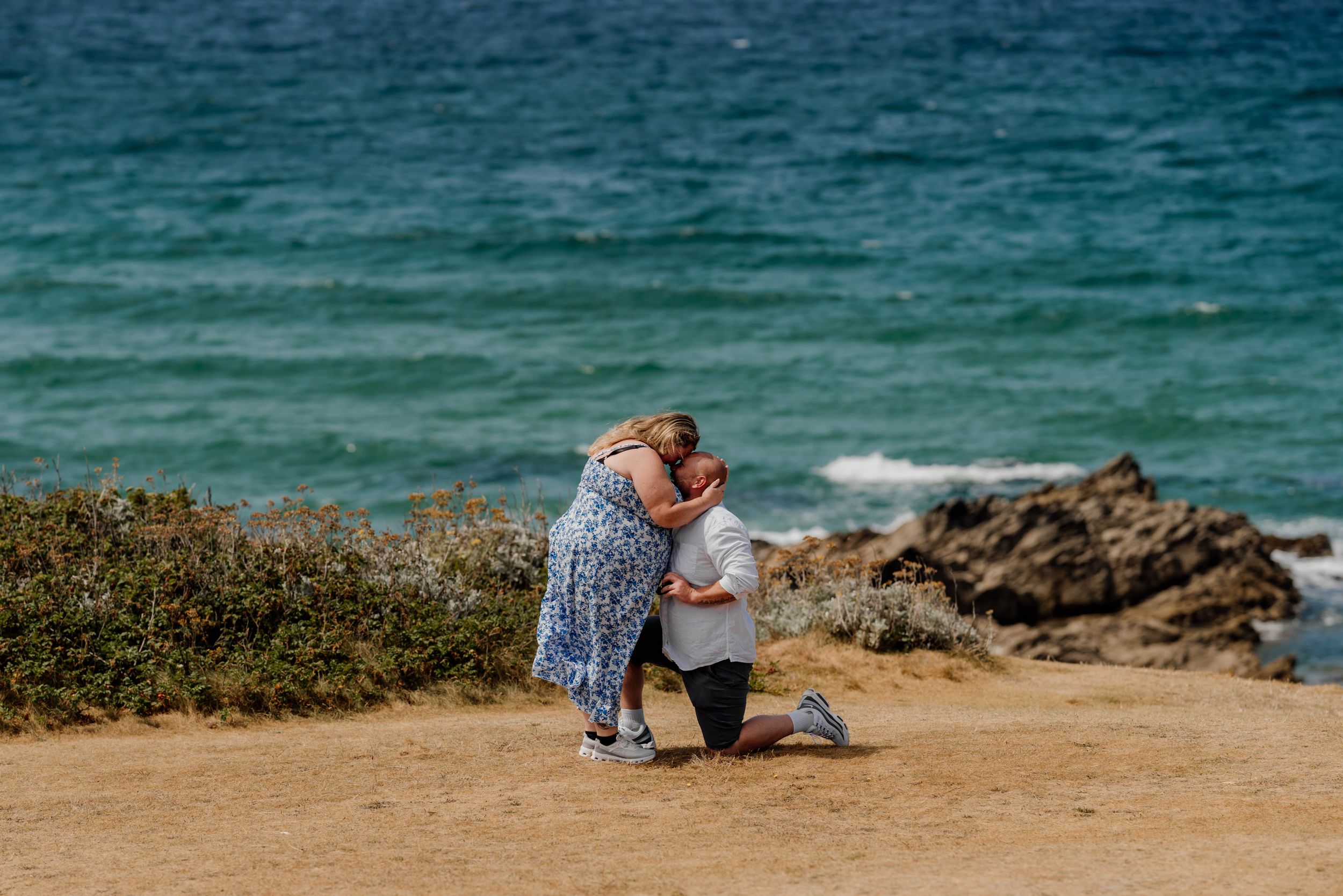 Couple embracing during a romantic beach proposal on a rocky coastal cliff with turquoise ocean waves in the background.