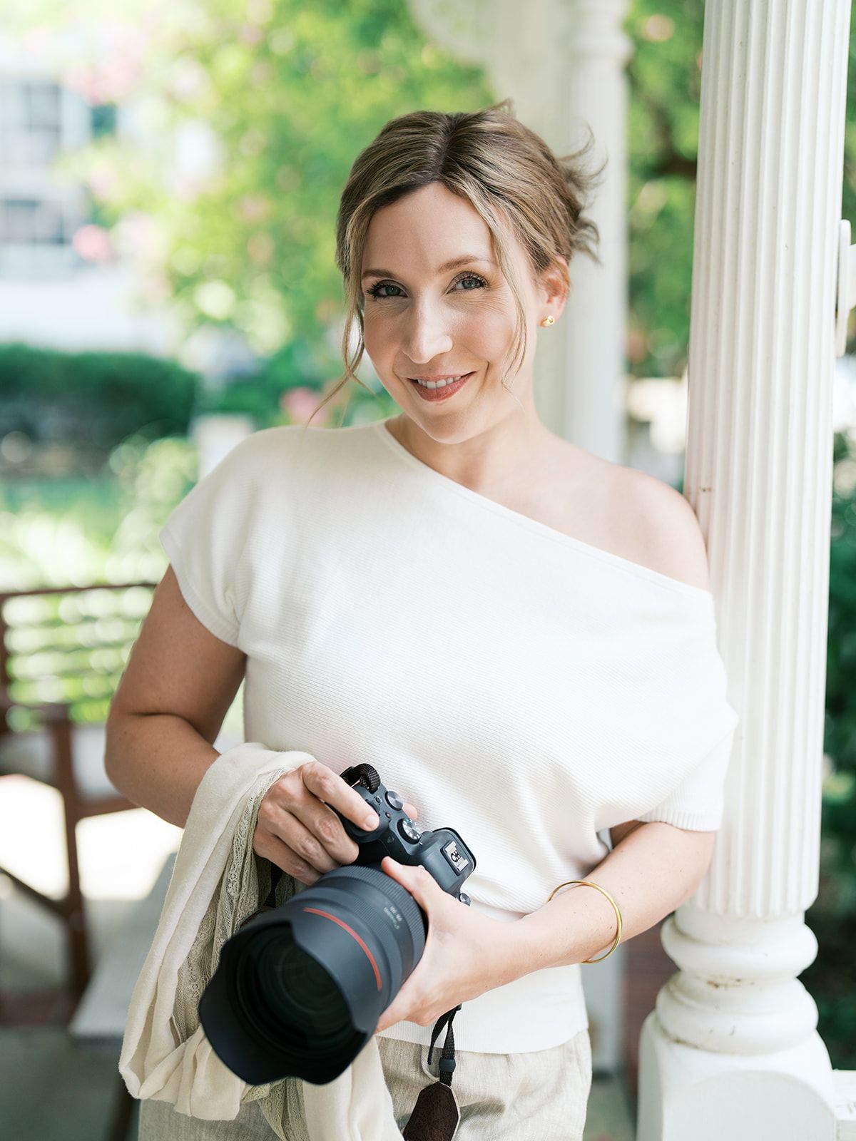 Smiling female photographer holding a Canon camera, wearing a white off-shoulder top, posing outdoors near white columns.