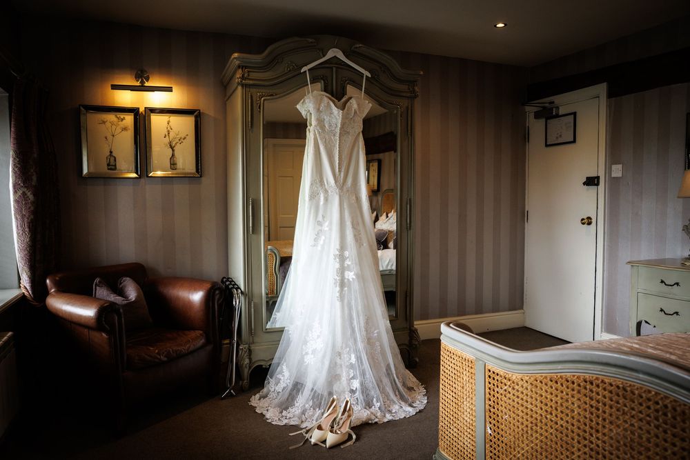 Elegant lace wedding dress hanging on a wardrobe in a warmly lit hotel bridal suite with leather armchair.