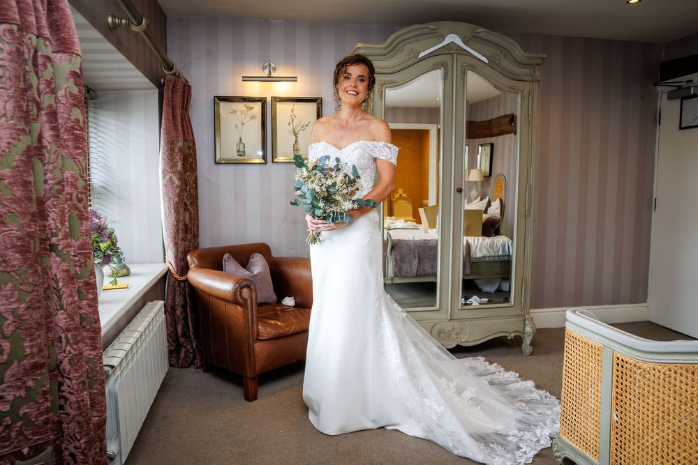 Bride in white off-shoulder gown holding bouquet stands in elegant hotel room with armchair and wardrobe.