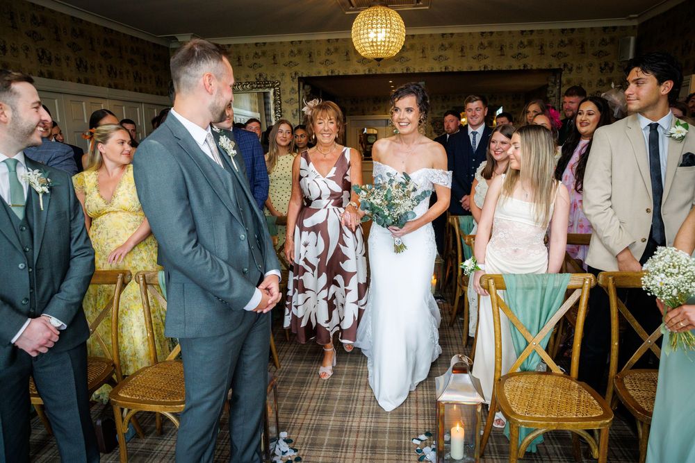 Bride walks down the aisle in white off-shoulder gown holding greenery bouquet at an elegant indoor wedding ceremony.