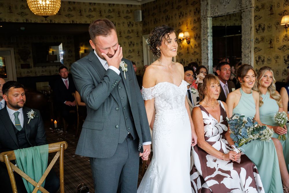 Emotional groom wiping tears beside smiling bride in white dress during wedding ceremony with guests seated behind.