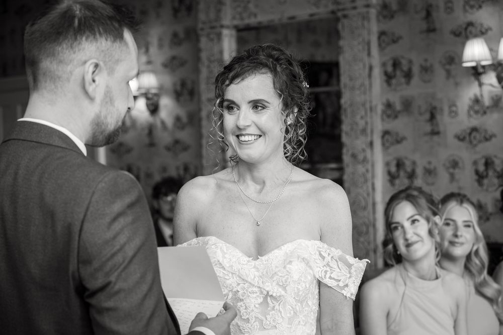 Black and white photo of a smiling bride in lace dress exchanging vows with groom at wedding ceremony.