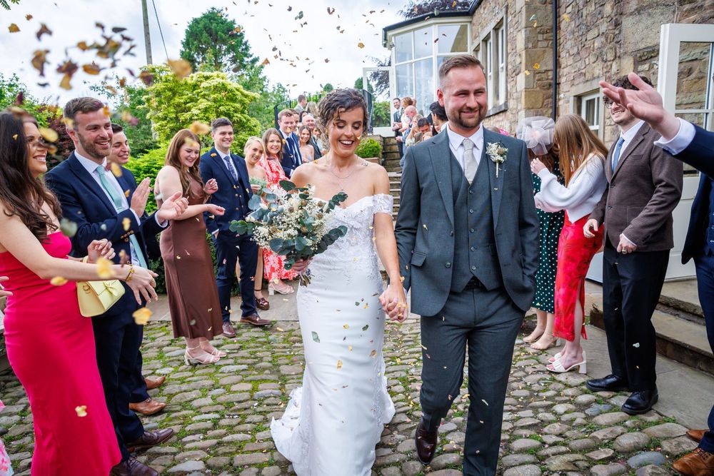 Bride and groom walk through confetti shower surrounded by cheering wedding guests on cobblestone path.