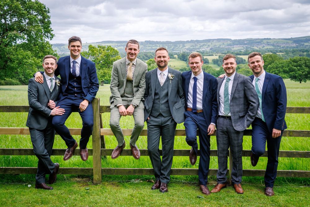 Seven groomsmen in suits posing by a wooden fence in a lush green countryside setting on a wedding day.
