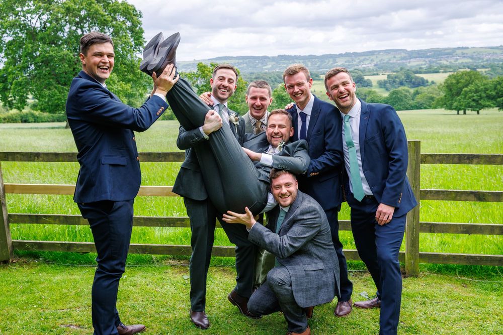 Groomsmen in navy suits lifting groom outdoors at countryside wedding venue with green fields backdrop.