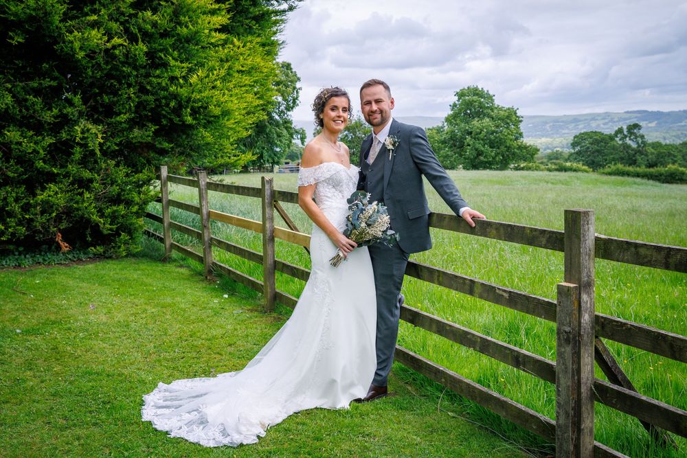 Bride and groom posing by rustic wooden fence in lush green countryside on their wedding day.