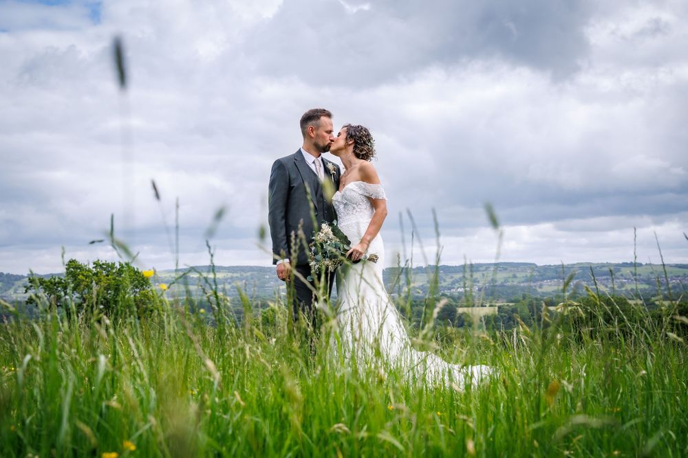 A bride and groom share a romantic kiss in a lush green meadow with a scenic countryside backdrop on their wedding day.