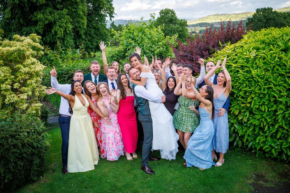 Joyful wedding party celebrating outdoors in garden with bride groom and guests cheering with arms raised.