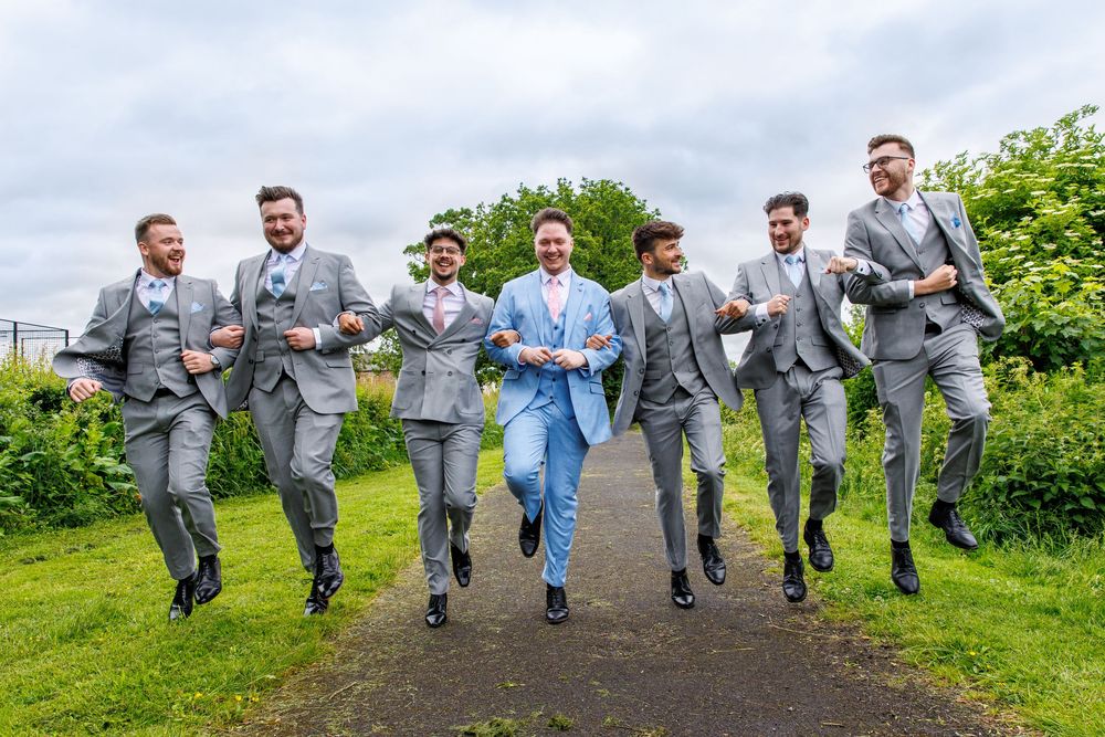 Groom in light blue suit walks confidently with six groomsmen in matching grey suits on an outdoor path.