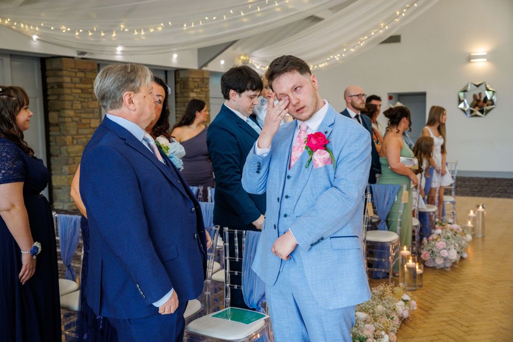Emotional groom in light blue suit wiping tears at wedding ceremony aisle, with guests standing in decorated venue.