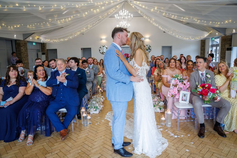 Bride and groom share first kiss at wedding ceremony with guests applauding in decorated venue with fairy lights.