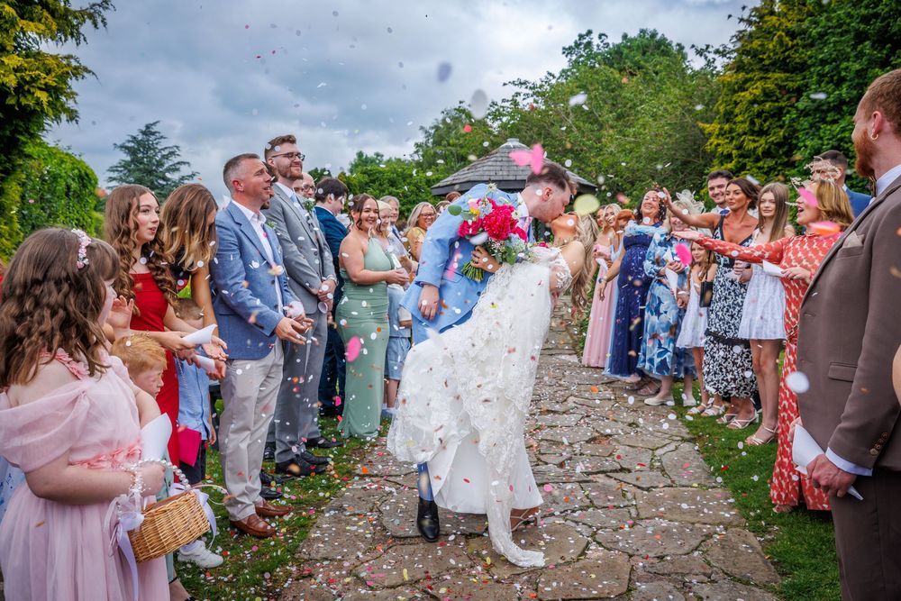 Newlyweds share a kiss while guests throw confetti along a garden pathway at an outdoor wedding celebration.