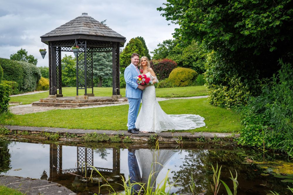 Bride and groom pose on a stone bridge beside a reflective pond with a gazebo and lush gardens at their wedding venue.
