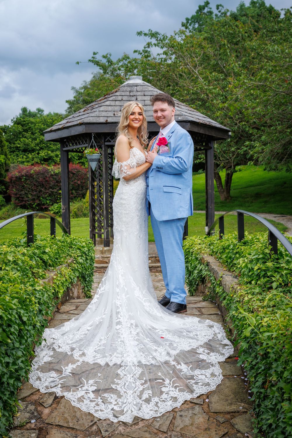 Bride in lace gown and groom in blue suit pose by garden gazebo on stone path.