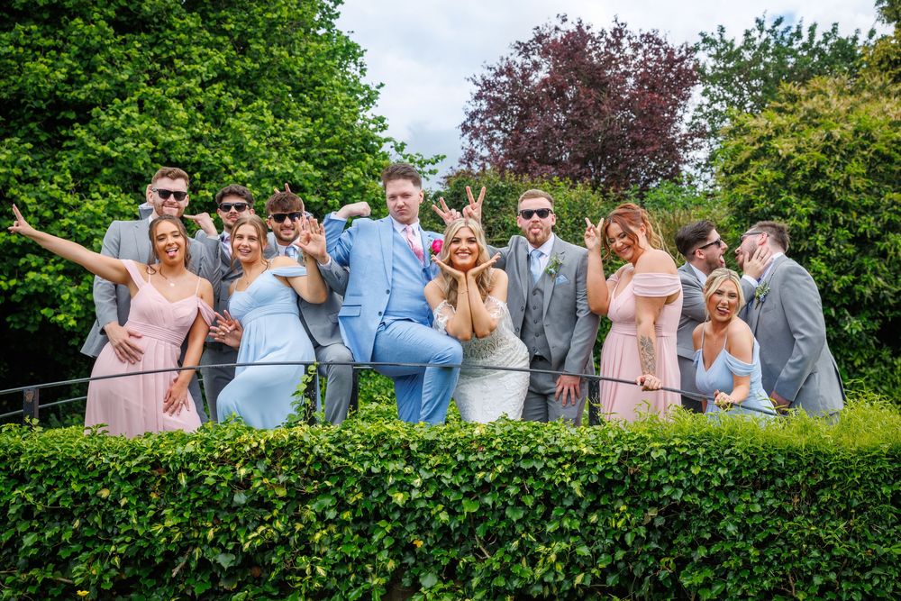 Wedding party posing playfully on a garden wall, wearing pink bridesmaid dresses and gray suits on a sunny day.