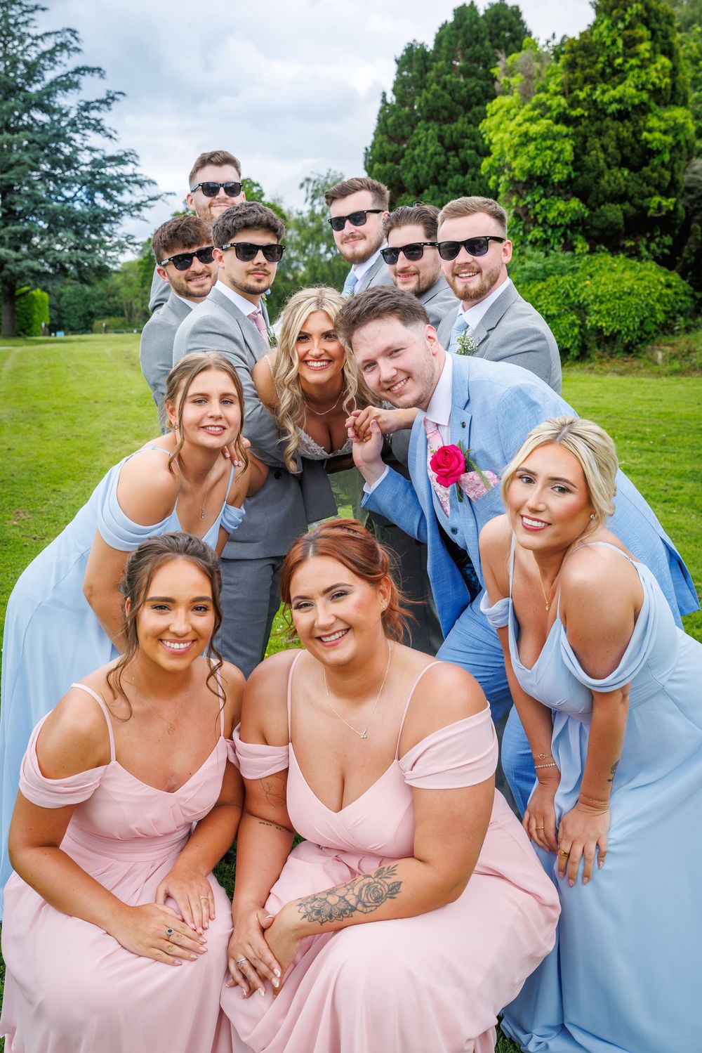 Wedding party poses outdoors, bridesmaids in blue and pink gowns with groomsmen in gray suits on lush green lawn.
