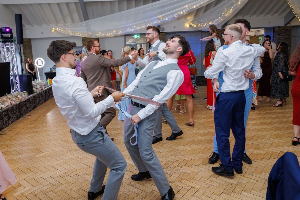 Men dancing energetically on a wooden dance floor at a wedding reception with guests in formal attire.