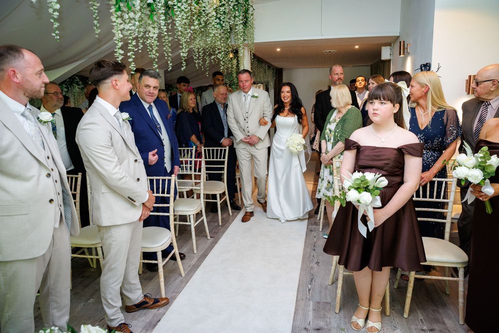 Bridesmaid in brown dress walks down aisle at elegant indoor wedding ceremony with guests seated on chiavari chairs.