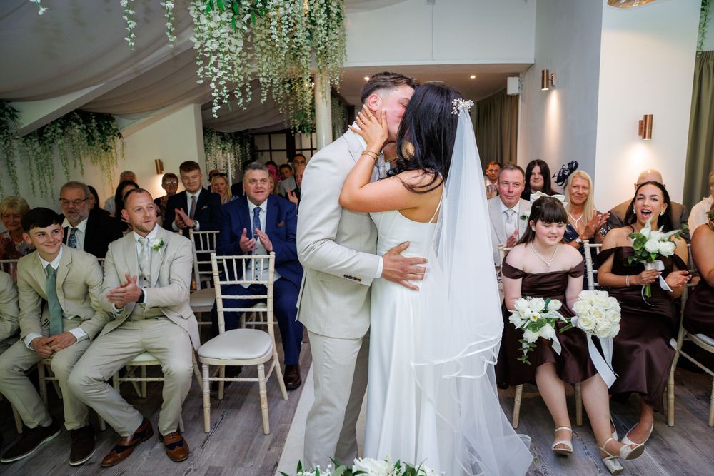 Bride and groom share first kiss at wedding ceremony while guests cheer in elegant white venue with floral decor.