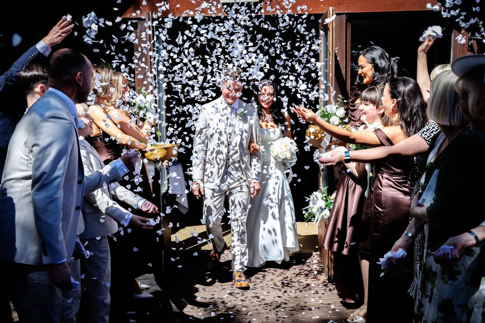 Newlywed couple walks through confetti shower surrounded by cheering guests at outdoor wedding ceremony.