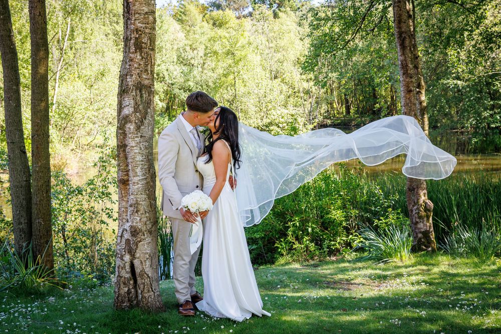 Bride and groom kissing in lush green garden, wedding veil flowing in wind, surrounded by trees near pond.
