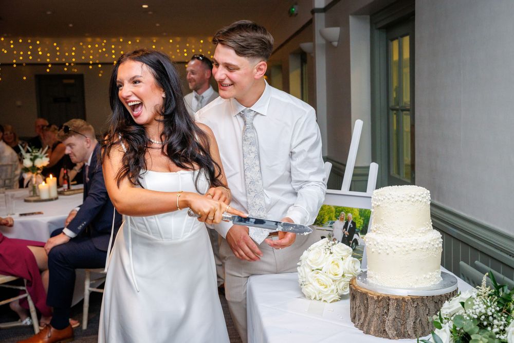 Bride and groom laughing while cutting white wedding cake at elegant indoor reception venue.